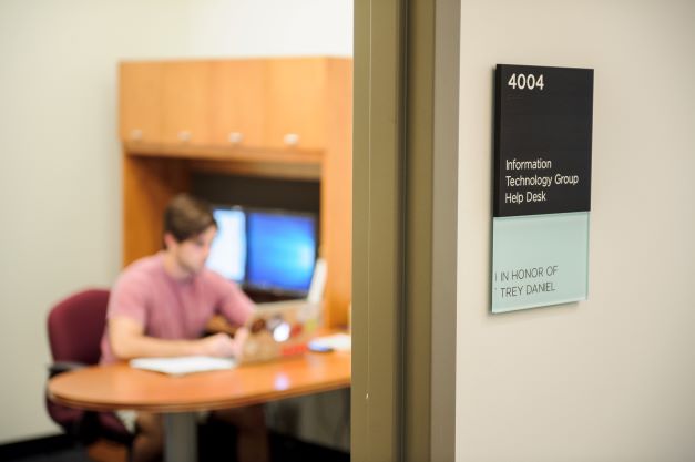 Person sitting a desk looking at a laptop screen. In the foreground there is an office sign that says ITG Helpdesk.
