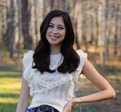 Ashlyn Puckett wears an ivory blouse with a lace and ruffle detail. The photo is taken outdoors.