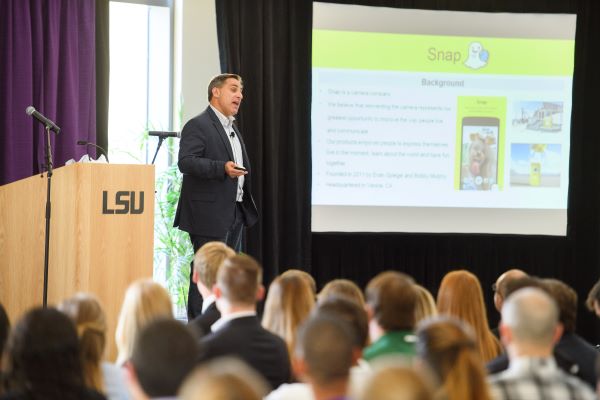 Man on stage with projector screen in background.